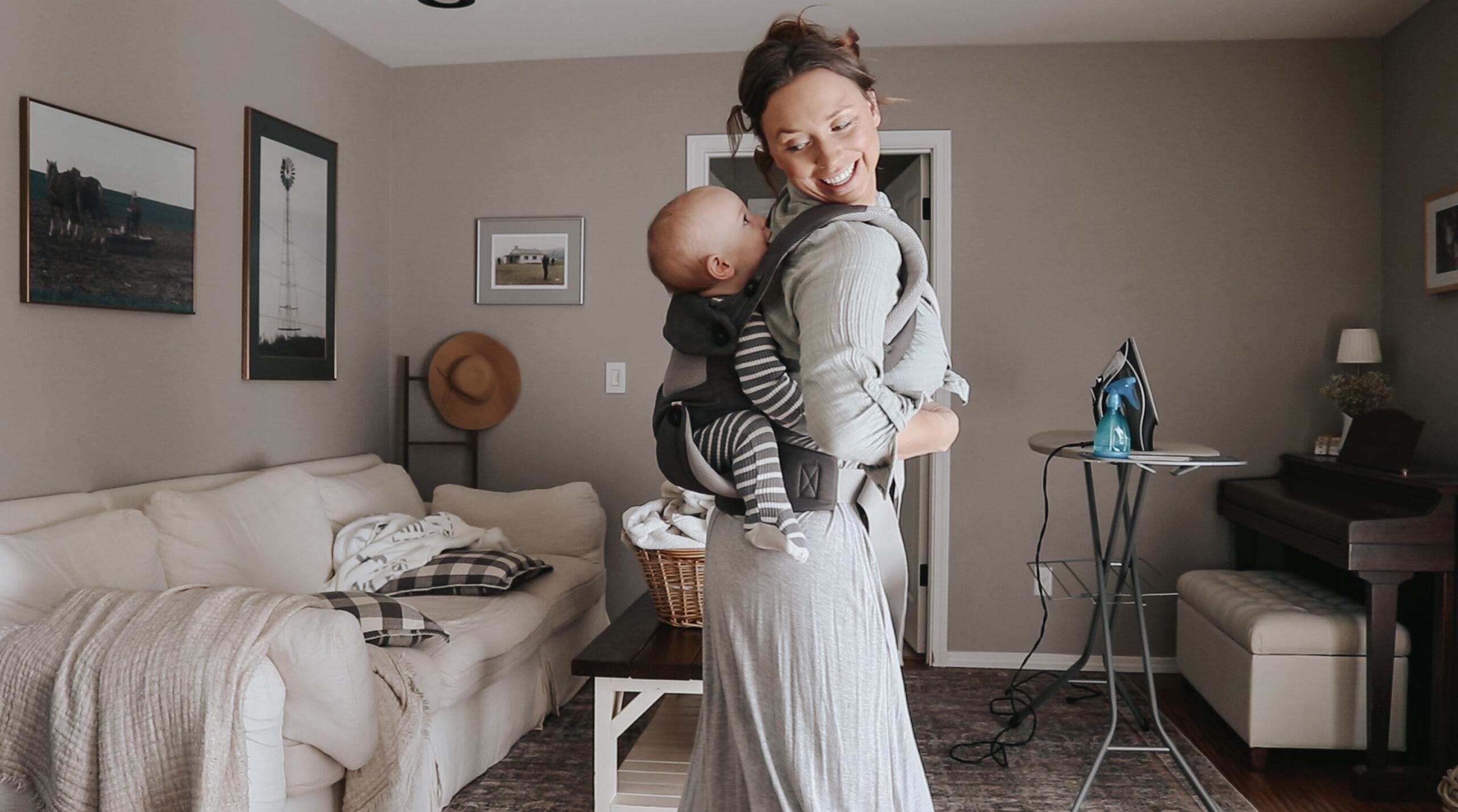 Christian mother puts baby in a carrier on her back. She is in the living room and there is a laundry basket on the coffee table with an ironing board next to it.